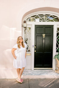 a woman in a white dress stands by a black door in front of a pink house in Charleston South Carolina