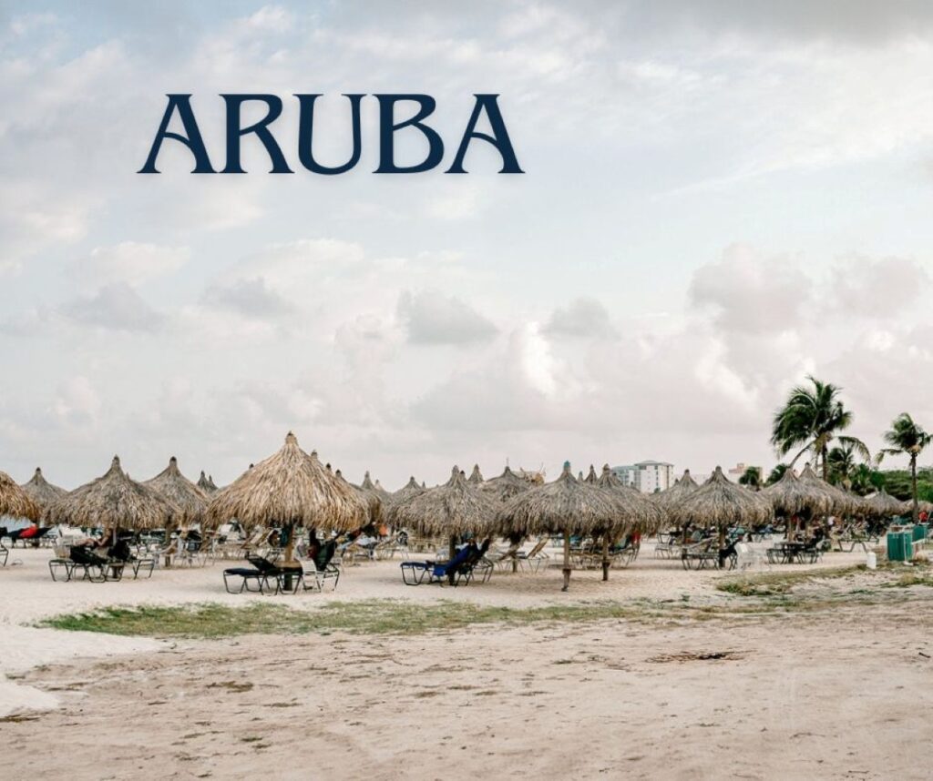 View of Eagle Beach in Aruba featuring rows of palapa-style thatched umbrellas and lounge chairs on white sand, with palm trees and a few buildings visible in the background under a partly cloudy sky.