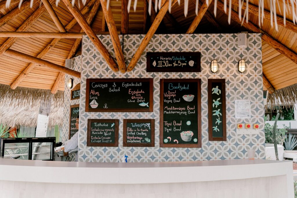Tropical outdoor food stand at Hyatt Ziva Puerto Vallarta featuring a thatched roof, decorative tiled wall, and chalkboard menus offering ceviche, salads, wraps, tostadas, and fresh bowls like vegan, Mediterranean, and Thai options.