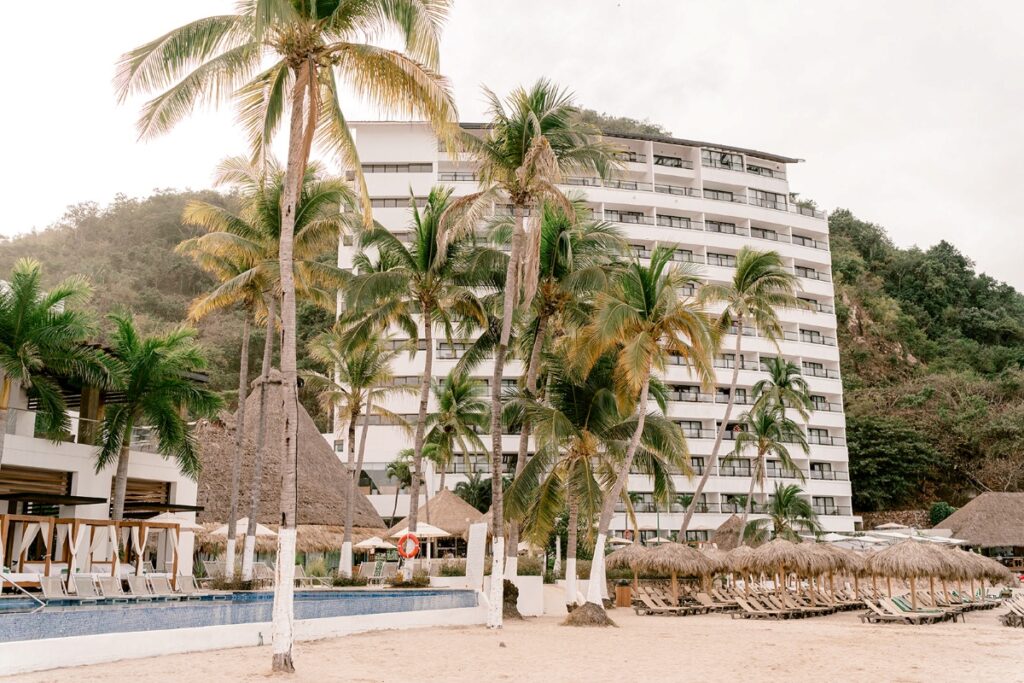 A tropical beachfront resort with a modern high-rise building, surrounded by lush palm trees, thatched-roof cabanas, and lounge chairs on the sandy beach, with an infinity pool in the foreground. This is the Hyatt Ziva Puerto Vallarta. 