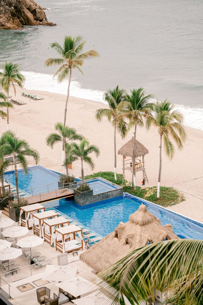 Scenic view of a tropical beachfront resort in Puerto Vallarta, Mexico, featuring palm trees, an infinity pool, and a sandy beach overlooking the Pacific Ocean with lush mountains in the background.