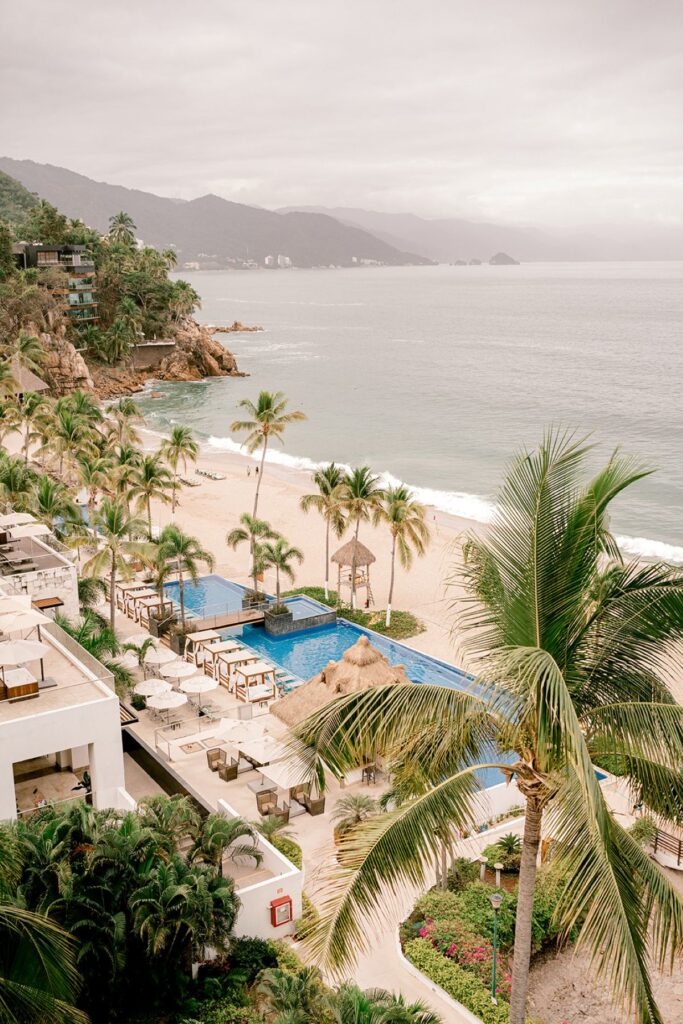 Scenic view of a tropical beachfront resort in Puerto Vallarta, Mexico, featuring palm trees, an infinity pool, and a sandy beach overlooking the Pacific Ocean with lush mountains in the background.
