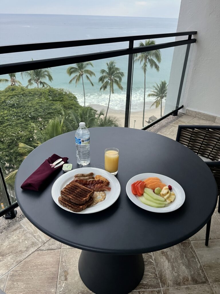 Room service breakfast on a balcony at Hyatt Ziva Puerto Vallarta, featuring a tropical ocean view with palm trees and sandy beach below. The table includes toast, eggs, bacon, sausage, fresh fruit, orange juice, and bottled water