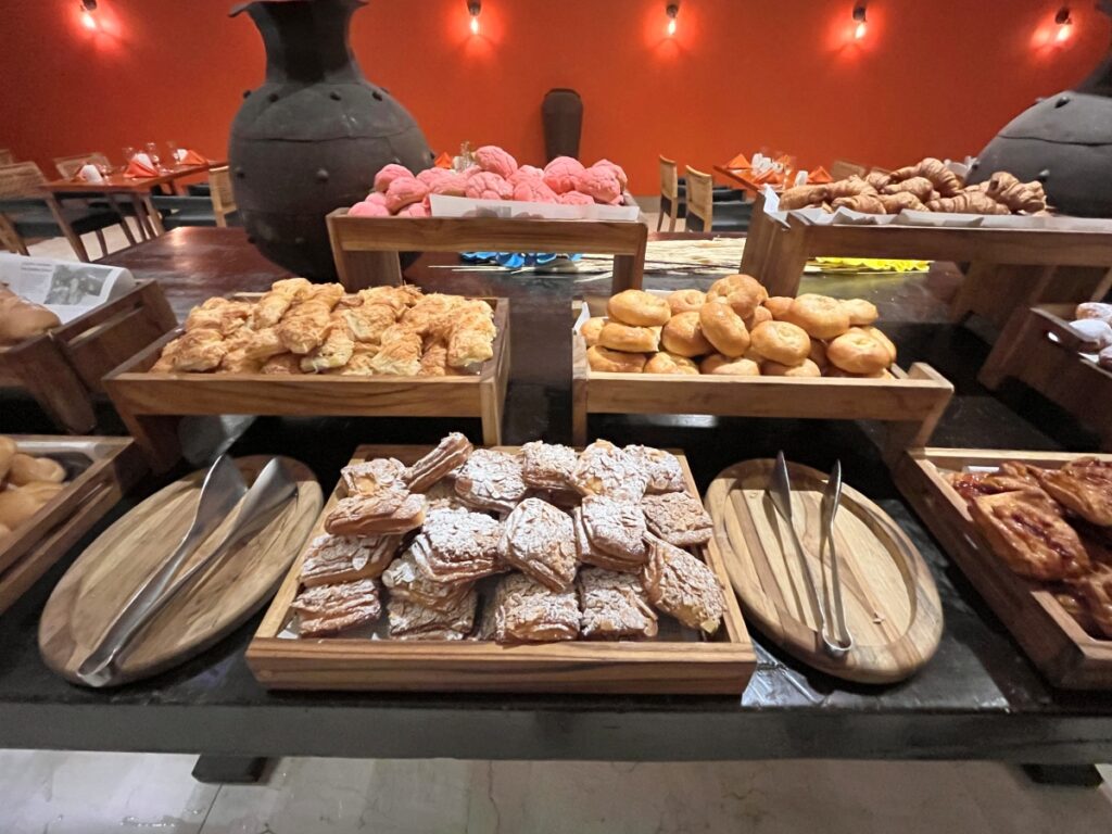 Buffet-style pastry and bread station, showcasing a variety of baked goods including croissants, conchas, puff pastries, and dinner rolls, displayed in wooden trays with serving tongs.