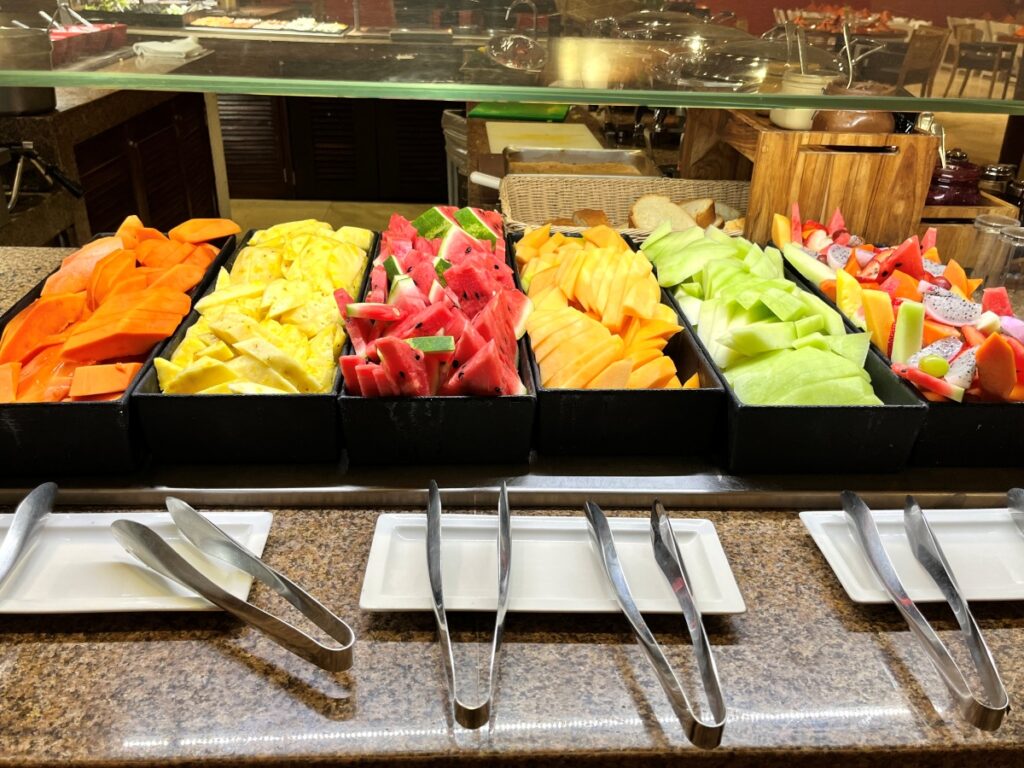 Colorful fruit display at the buffet, featuring neatly arranged trays of papaya, pineapple, watermelon, cantaloupe, honeydew, and dragon fruit, with serving tongs and plates ready for guests.