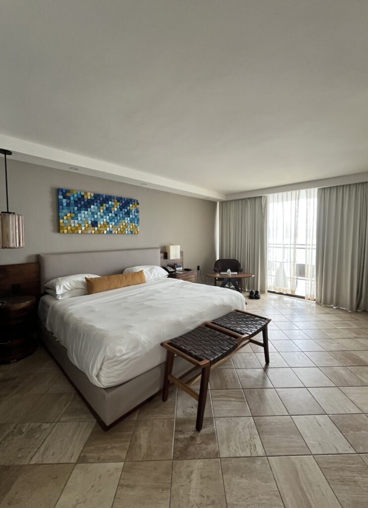 Spacious guest room at Hyatt Ziva Puerto Vallarta featuring a large king bed with modern decor, tile flooring, cozy seating area, and floor-to-ceiling windows with sheer curtains leading to a private balcony.