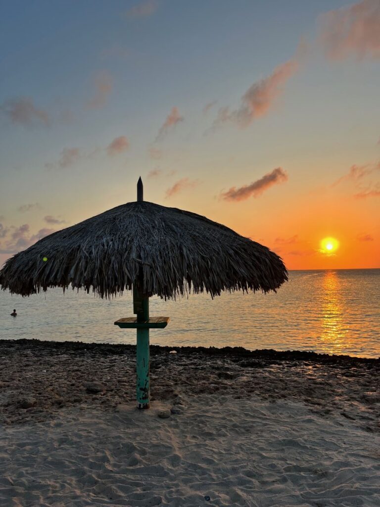 A serene sunset on the beach in Aruba, with a rustic palapa umbrella silhouetted against the glowing sky and golden sun reflecting off the calm ocean waves—capturing the tranquil beauty of island evenings.