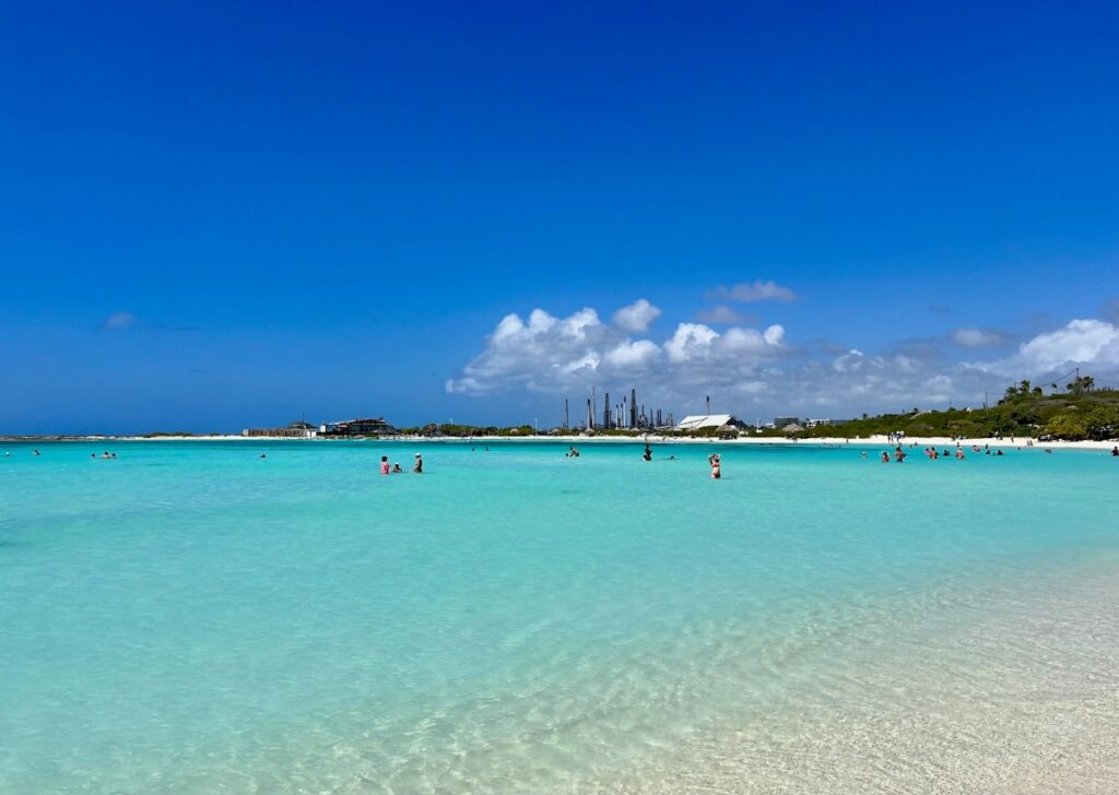 Visitors wading in the calm, shallow turquoise waters of Baby Beach in Aruba, with soft white sand underfoot and a backdrop of blue sky, scattered clouds, and a distant refinery on the horizon.