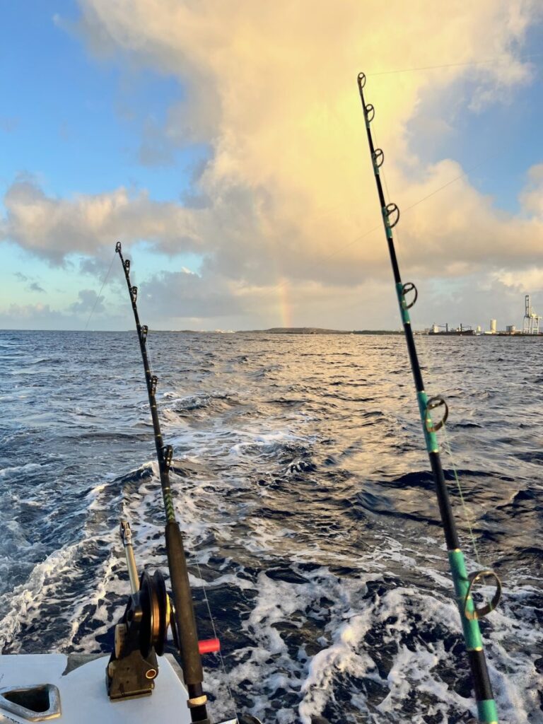 A view from the back of a fishing charter boat in Aruba, with fishing rods set against the open sea, foamy wake trailing behind, and a faint rainbow glowing beneath dramatic morning clouds near the island’s shoreline.