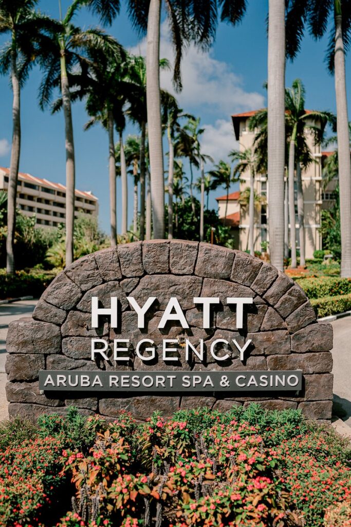 The stone entrance sign for the Hyatt Regency Aruba Resort Spa & Casino, surrounded by vibrant tropical flowers and tall palm trees, sets a luxurious tone as guests arrive at this upscale Caribbean getaway.