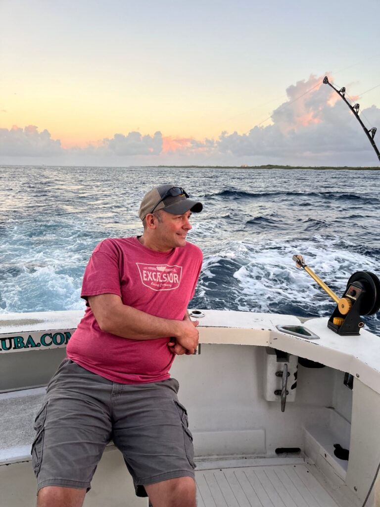 A man enjoying a sunrise fishing trip off the coast of Aruba, sitting relaxed on a charter boat with a fishing rod set and waves trailing behind, as soft morning light colors the sky with pastel hues.