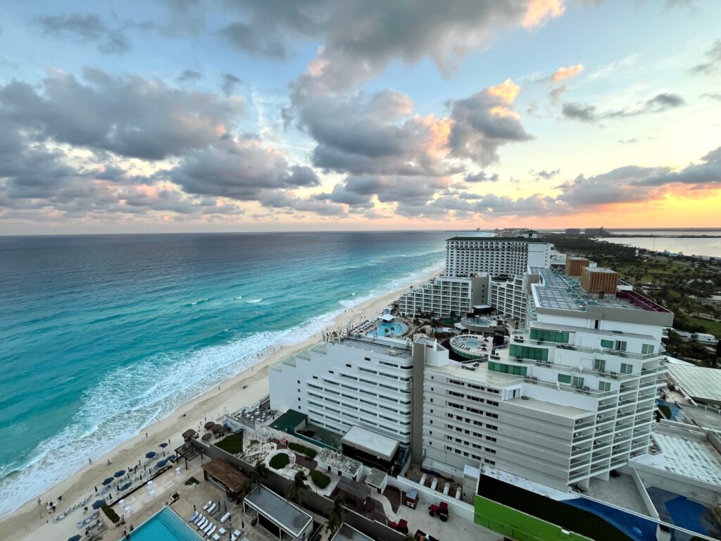 A high-rise balcony view at Secrets The Vine Cancun overlooking the turquoise Caribbean Sea, white-sand beach, and resort pools at sunset with soft clouds painted in pastel colors across the sky