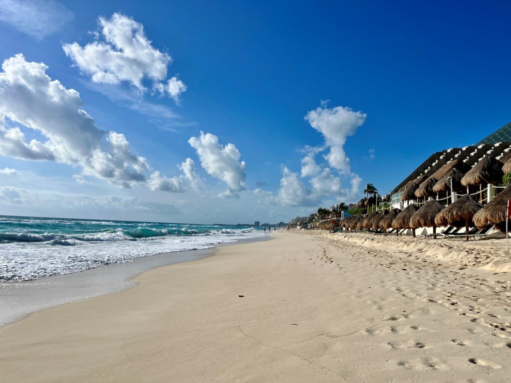 A sunny morning view along the white-sand beach at Secrets The Vine Cancun, with turquoise Caribbean waves rolling in and rows of thatched palapa umbrellas lining the shore under a bright blue sky with scattered clouds.