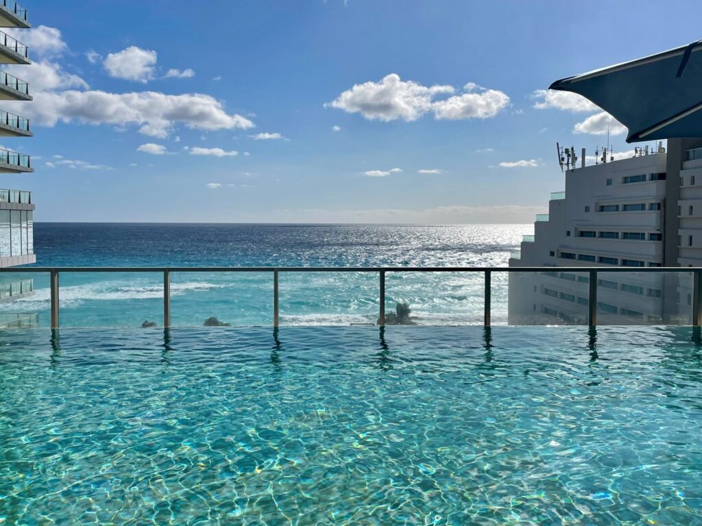 Sparkling infinity pool at the Preferred Club of Secrets The Vine Cancun overlooking the turquoise Caribbean Sea, with modern hotel buildings on either side and the sun glistening on the water under a clear blue sky