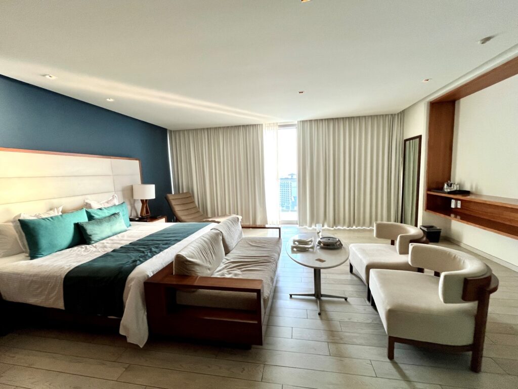 Spacious modern guest room at Secrets The Vine Cancun featuring a king-size bed with teal accents, a cozy seating area with armchairs and a sofa, and floor-to-ceiling windows with light curtains letting in natural light