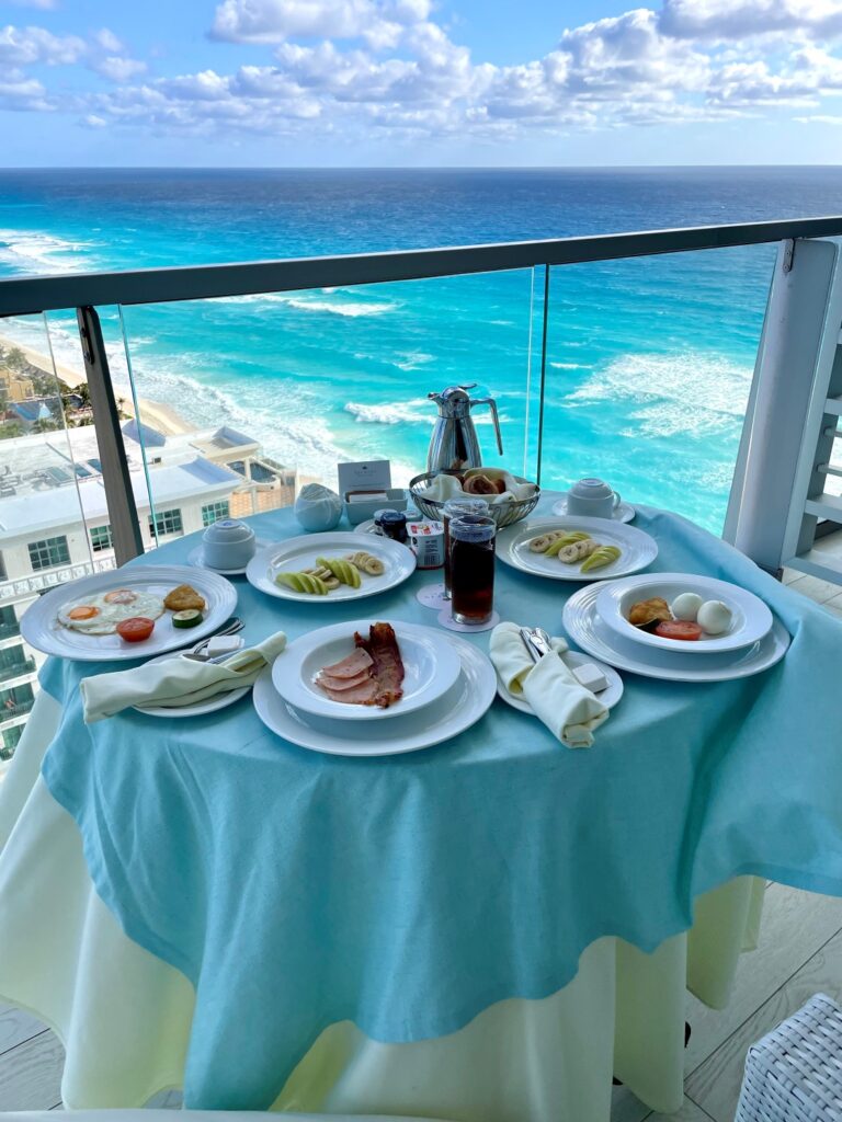 Room service breakfast set on a balcony at Secrets The Vine Cancun, featuring plates of eggs, bacon, fruit, and pastries on a turquoise tablecloth overlooking the turquoise Caribbean Sea and white sandy beach below