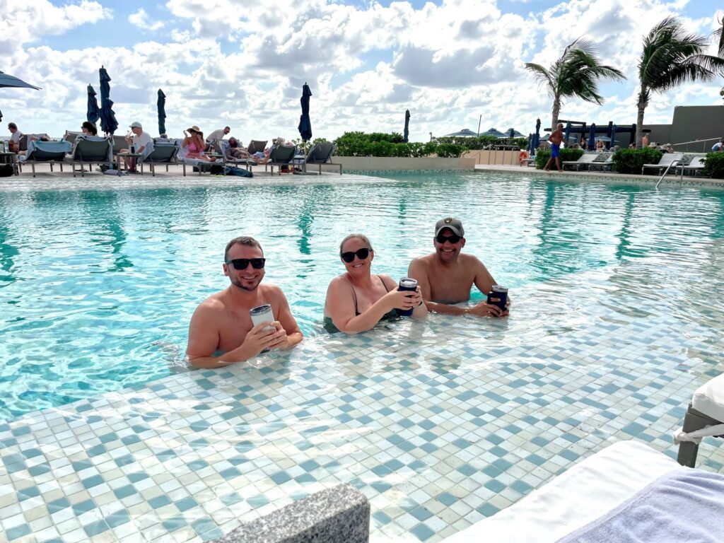Three adults smiling and holding drinks while standing in the infinity pool at Secrets The Vine Cancun, with palm trees, pool loungers, and ocean-side resort views behind them.