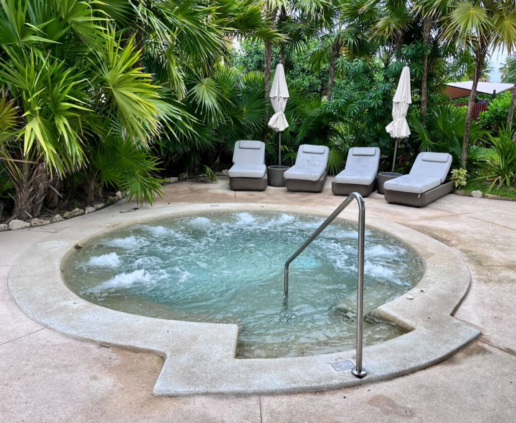 Secluded outdoor hot tub at Secrets Maroma Beach Riviera Cancun surrounded by lush tropical palms, bubbling water jets, and cushioned lounge chairs with umbrellas for relaxing in the Caribbean sunshine.