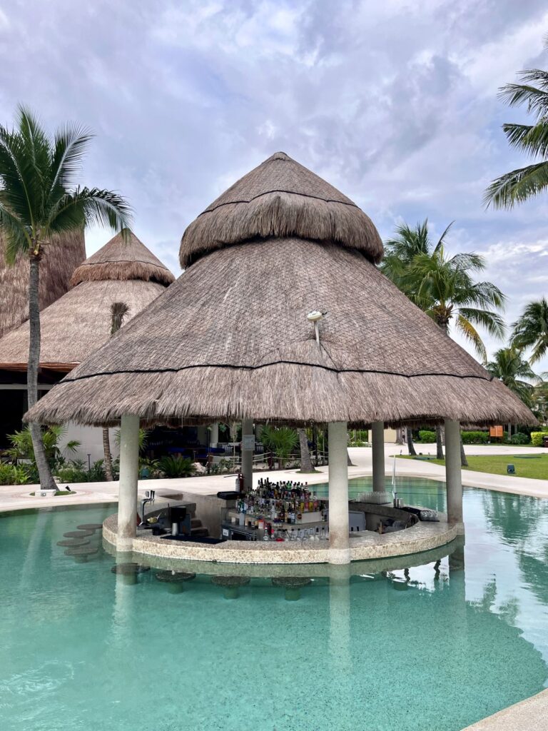 Swim-up pool bar at Secrets Maroma Beach Riviera Cancun featuring a circular thatched-roof palapa, turquoise water, and bar seating in a tropical adults-only resort setting.
