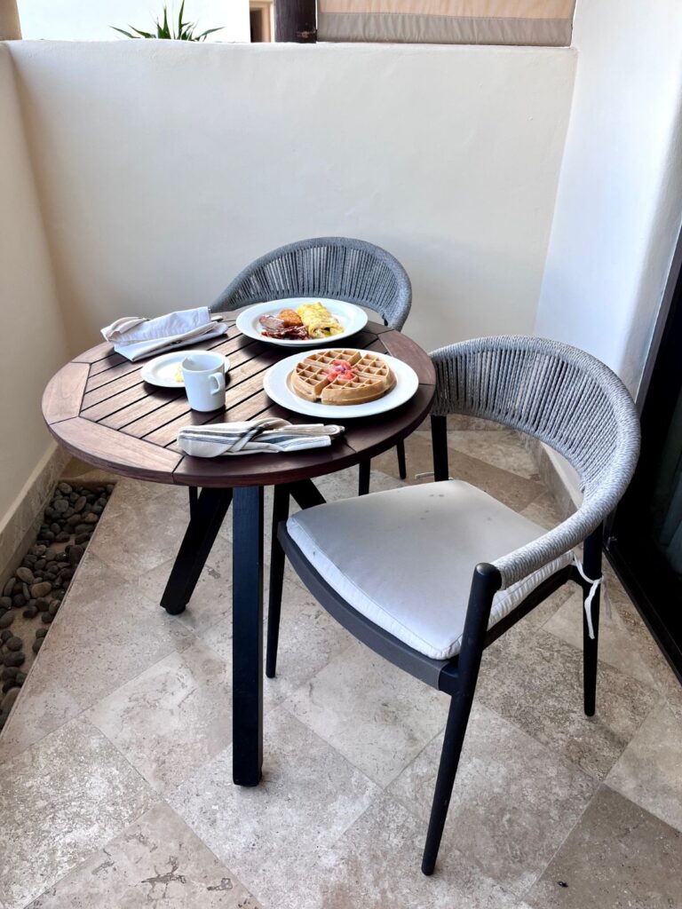Private balcony breakfast at Secrets Maroma Beach Riviera Cancun featuring a wooden bistro table with waffles topped with fresh fruit, an omelet plate, coffee, and cushioned woven chairs overlooking a serene resort setting