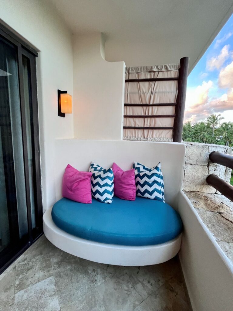 Cozy private balcony seating at Secrets Maroma Beach Riviera Cancun featuring a curved turquoise daybed with pink and blue chevron pillows, overlooking lush palm trees and a colorful Riviera Cancun sunset.